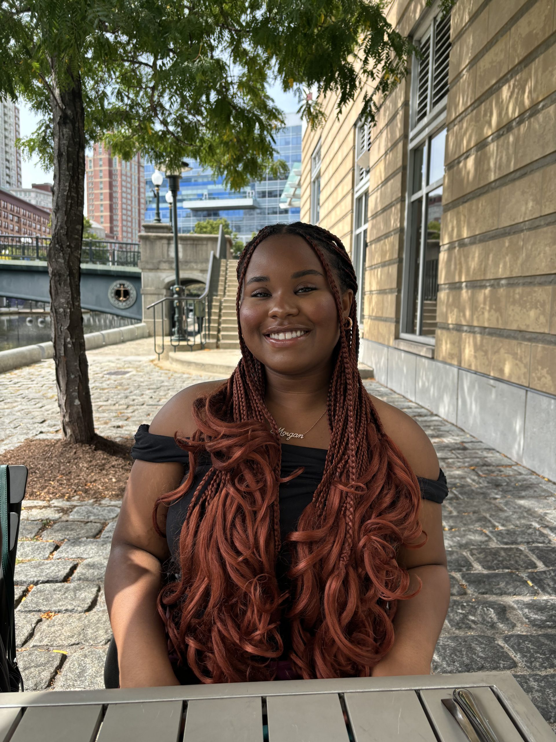 African American young woman with braided hair sitting down. Wearing a black off shoulder top