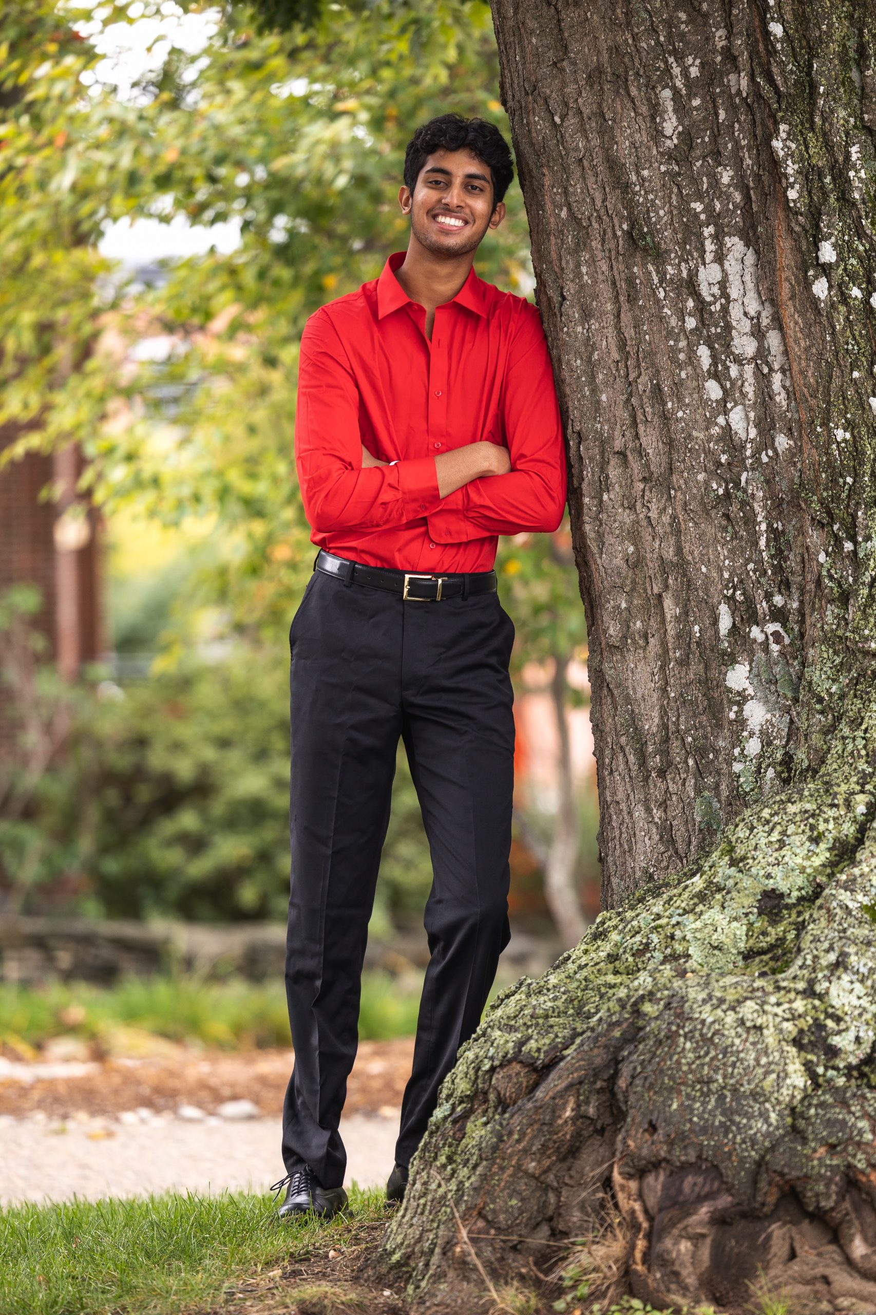 Keshav Desibhatla Male wearing red shirt and black pants. Photo in natural background. He is leaning against a tree.