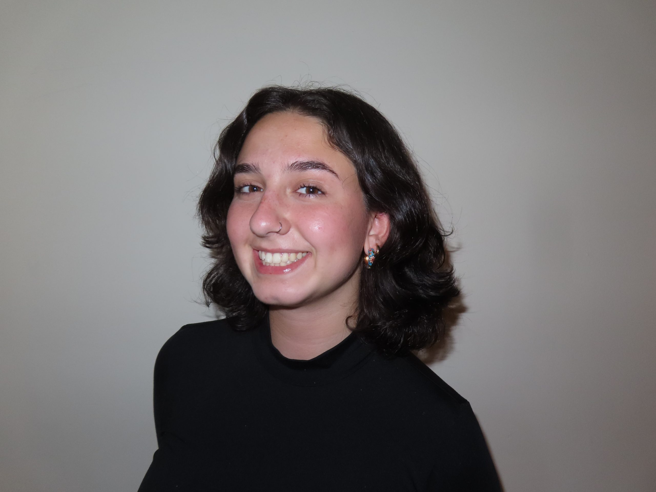 Young woman with dark brown hair. She has a big smile and is leaning against a gray background